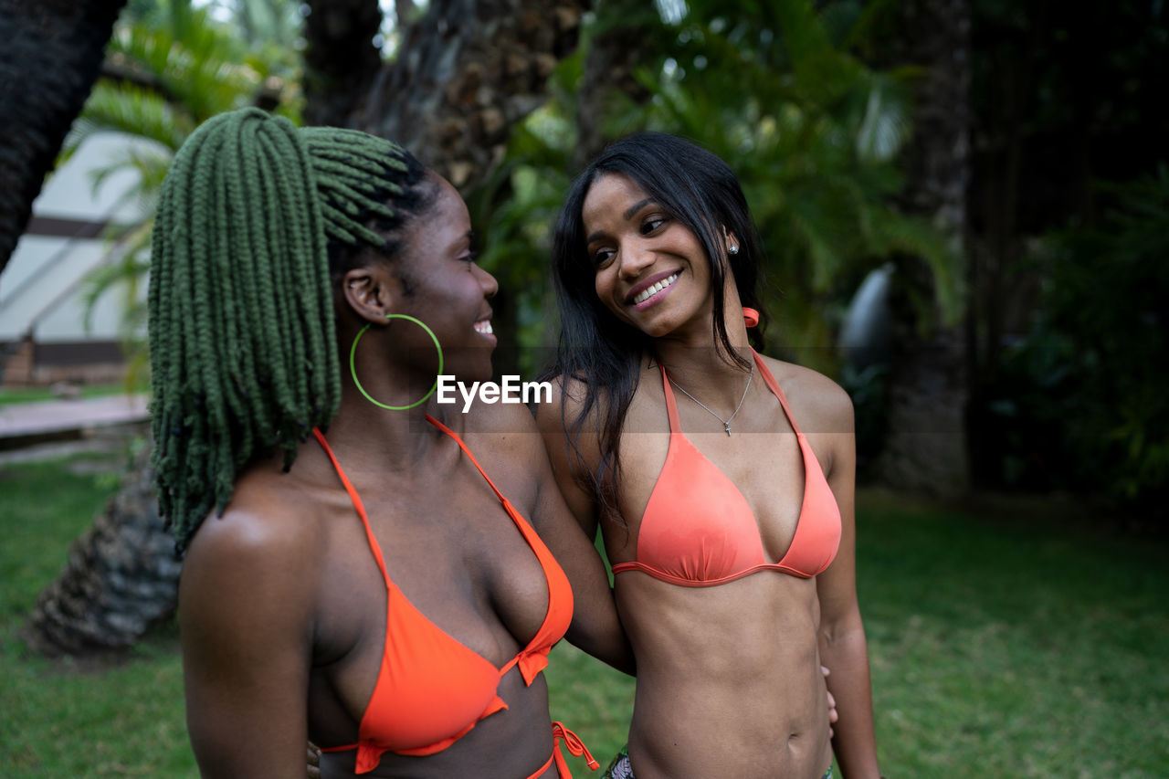 Happy african american female friends in bikinis looking at each other while standing on lawn in tropical country on summer day