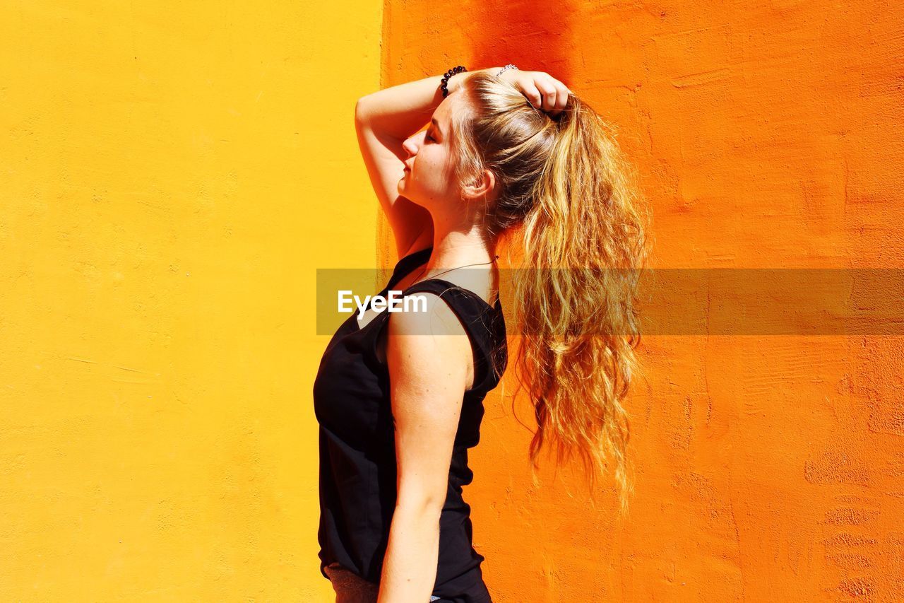 Side view of young woman with hand in hair standing against orange wall