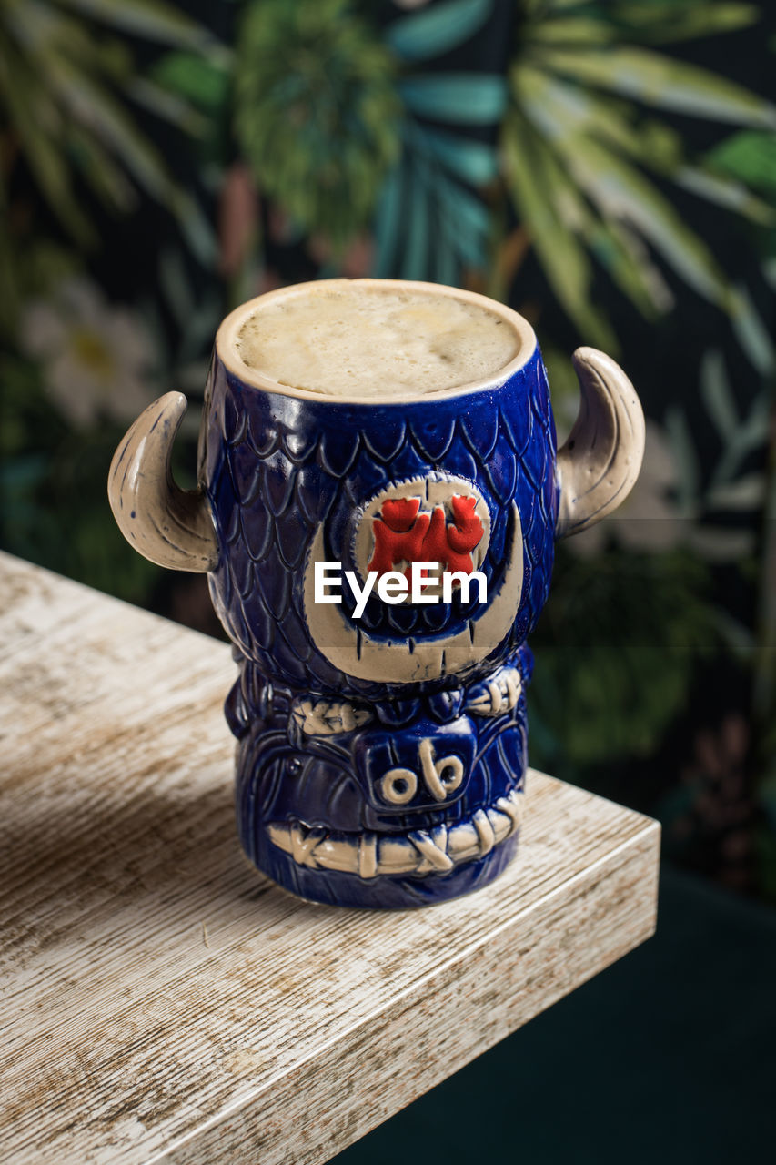 From above of bull shaped tiki mug of alcohol drink with froth placed against wooden table on blurred background