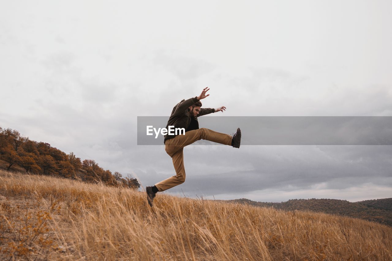 Full length of young man jumping on field against cloudy sky