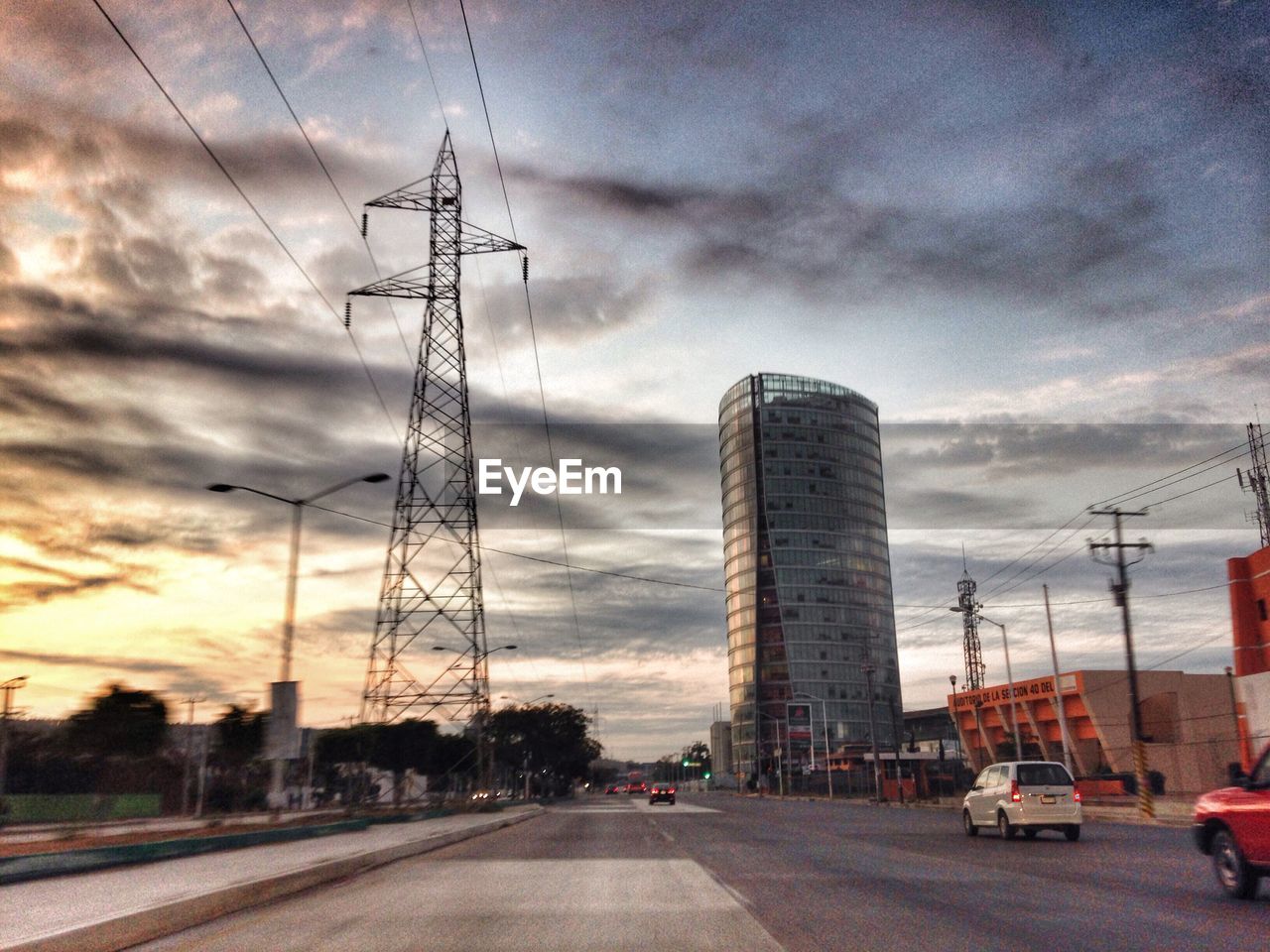 Road by modern building and electricity pylon against sky at sunset