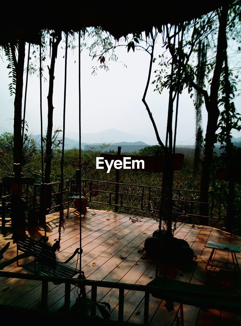 Empty swing on porch by trees against sky