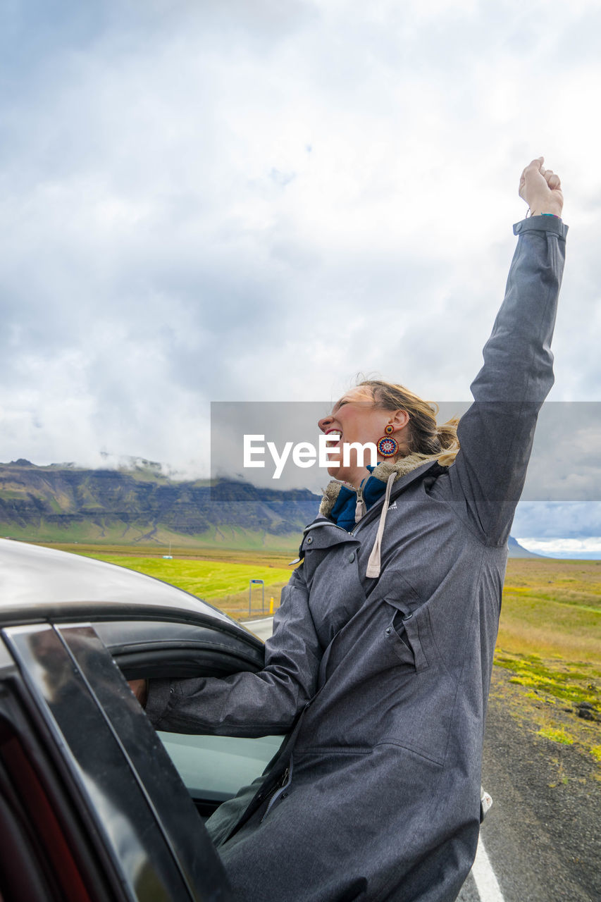 Happy mid adult woman with hand raised screaming while sitting on car window against cloudy sky