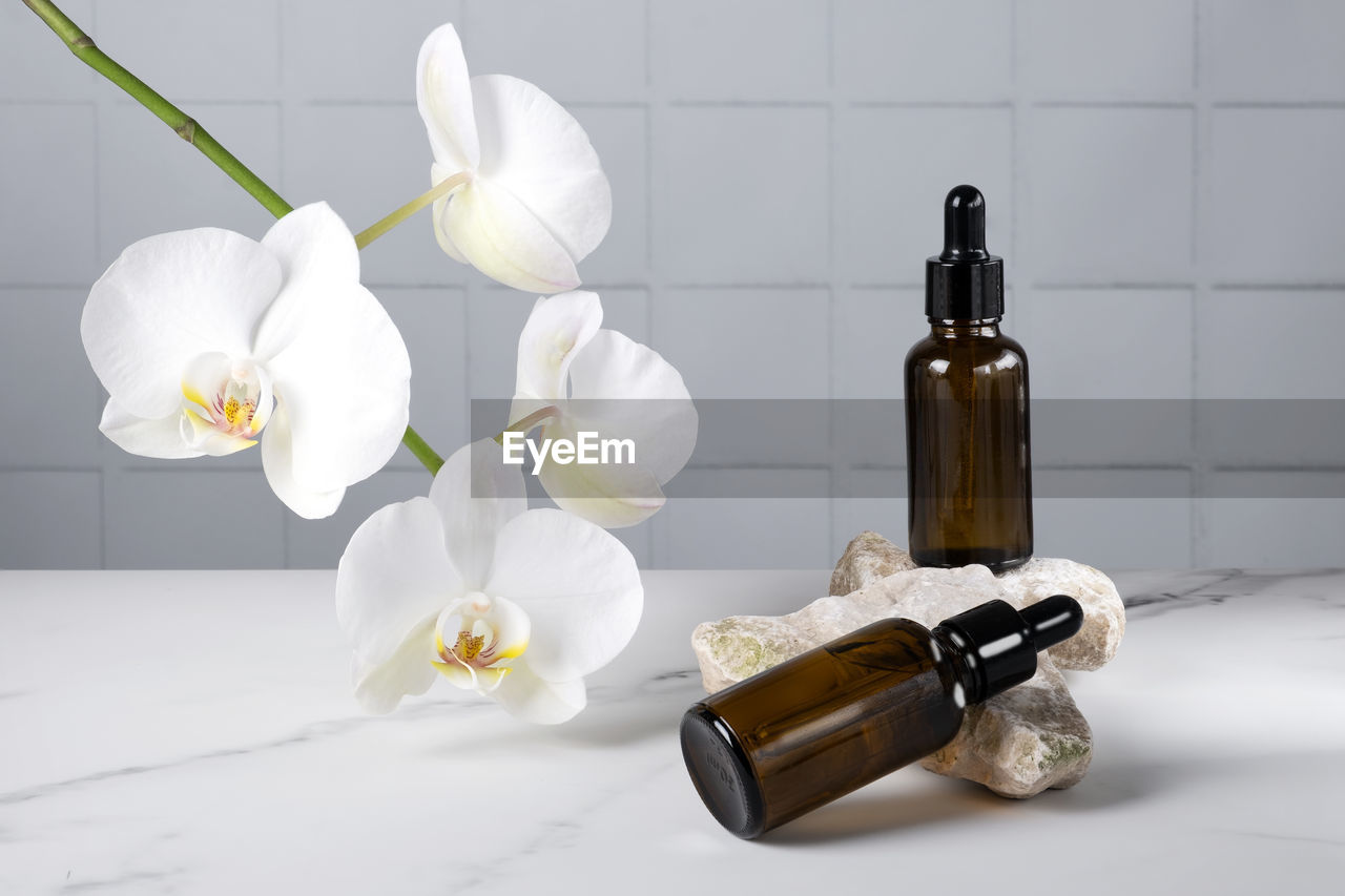 Bottles with pipette with cosmetic product for body skin care on stone against blooming orchid