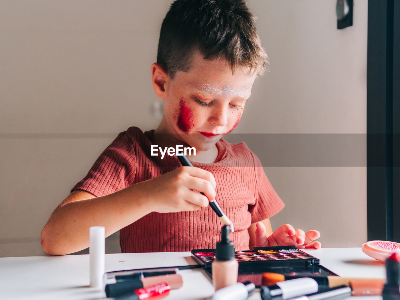 Charming child with makeup applicator looking away at table with eyeshadow palette