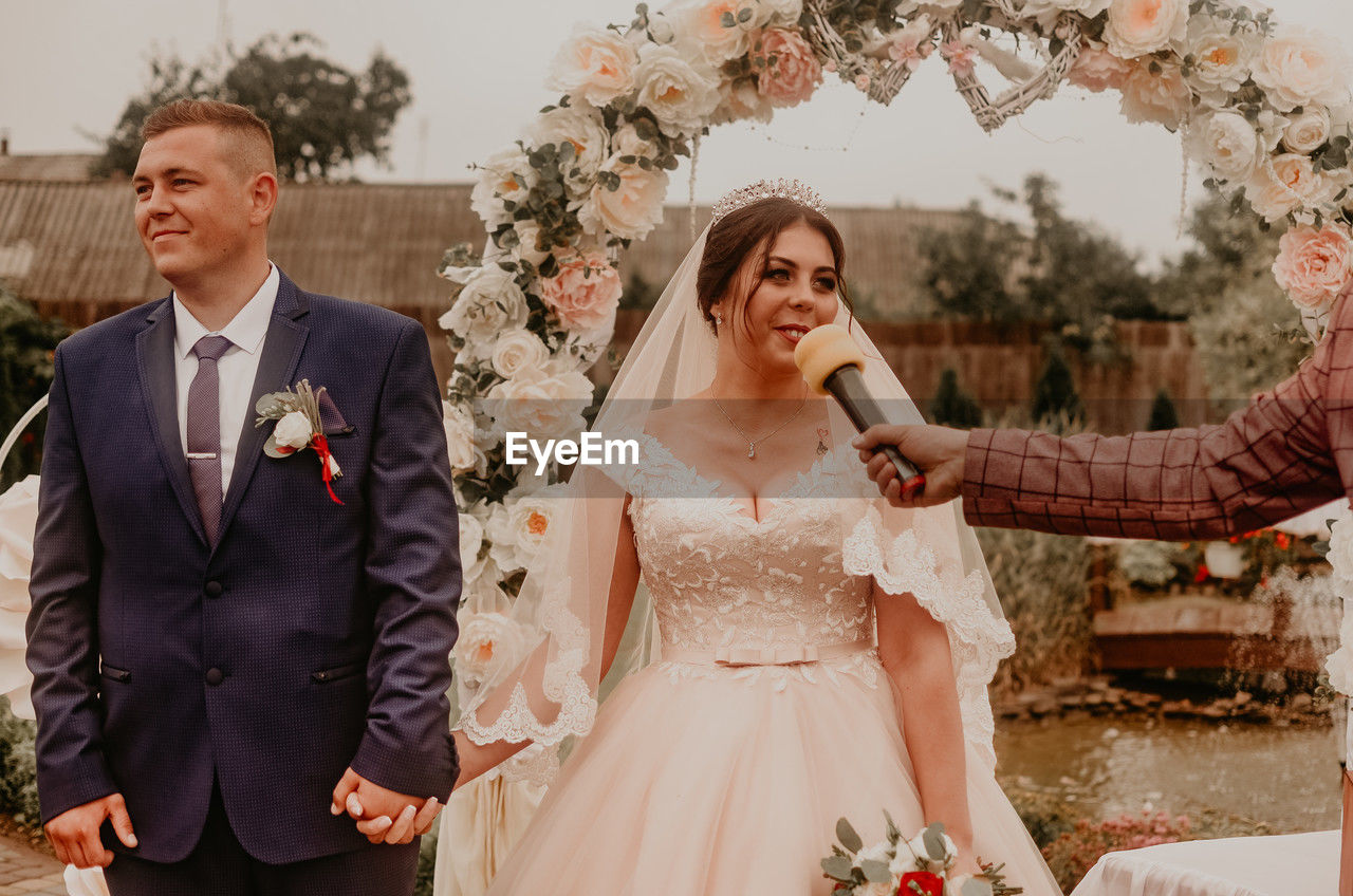 portrait of bride and bridegroom holding bouquet while standing against trees