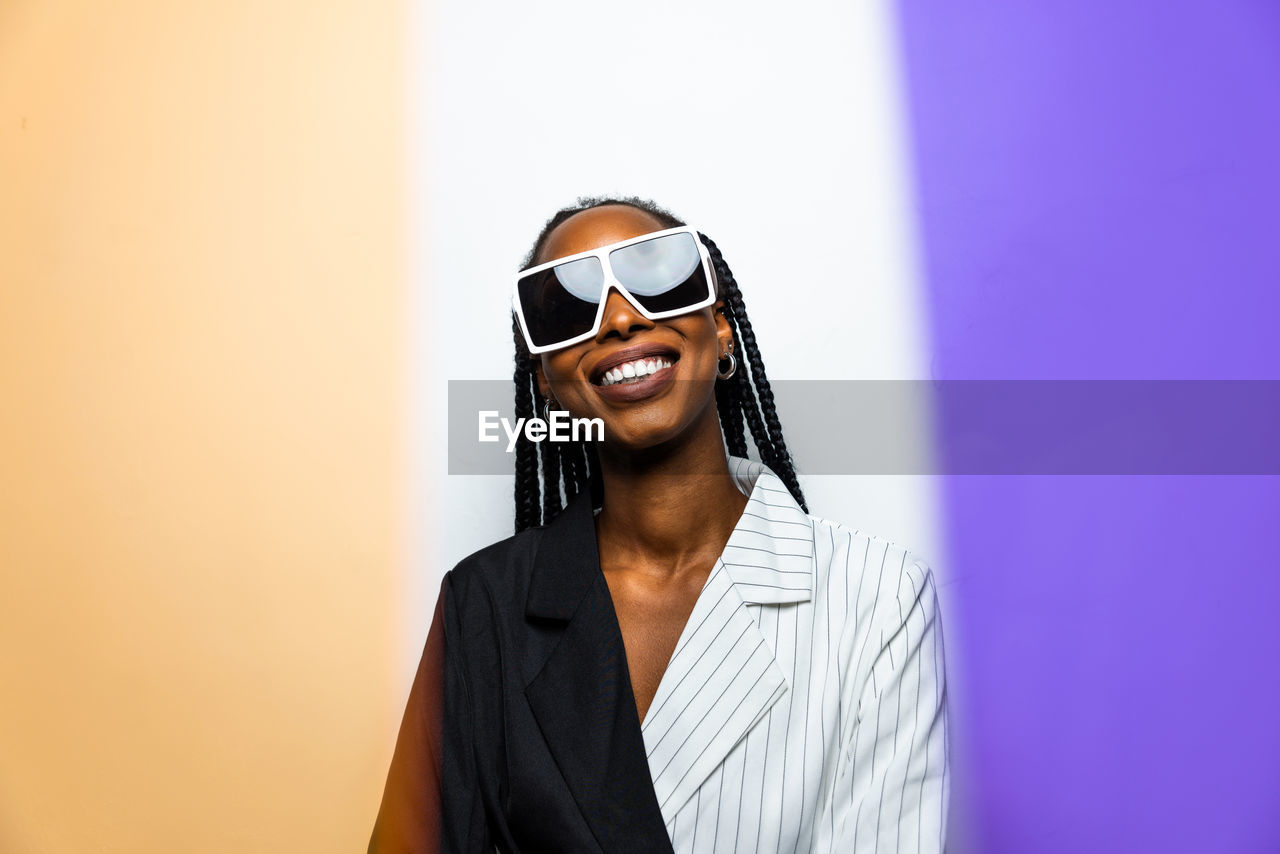 Portrait of smiling young woman wearing sunglasses standing against wall
