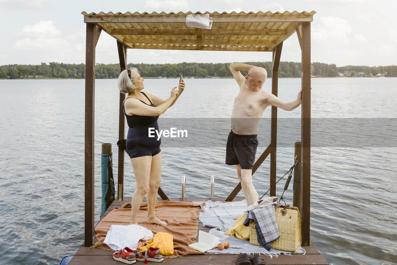 Full length of senior woman photographing shirtless man while standing in gazebo against river