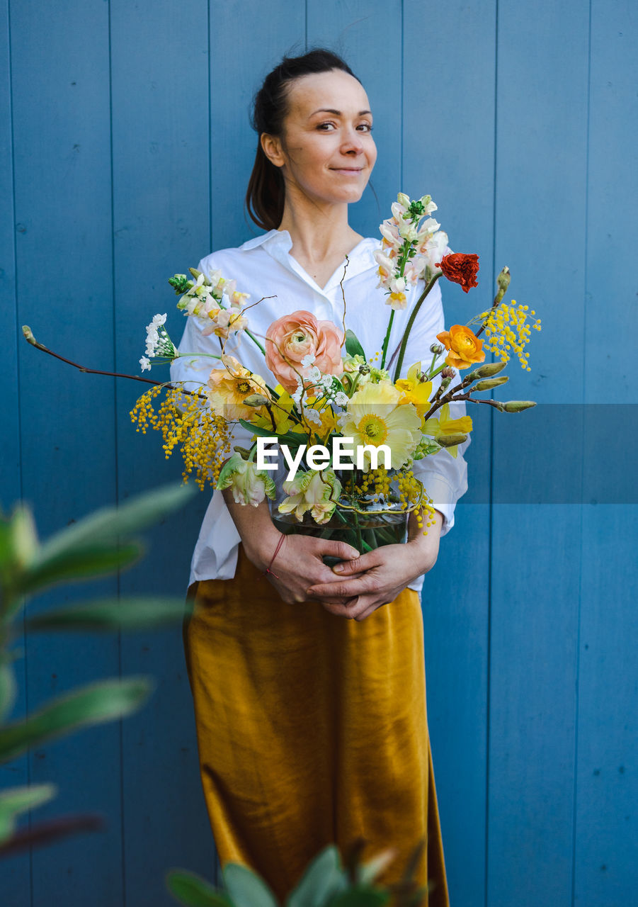 Young woman in flowing yellow skirt and white shirt with beautiful bouquet of peonies in glass vase