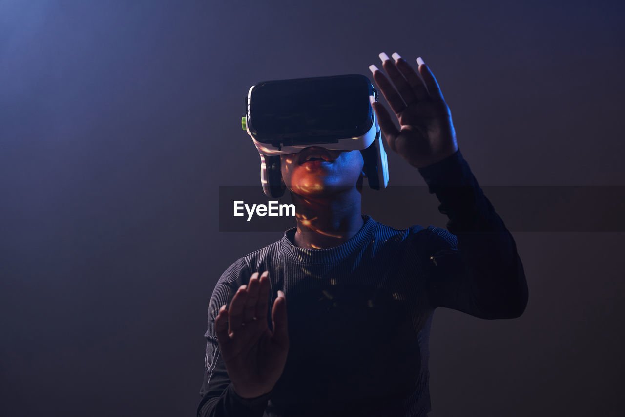 African american female in modern vr headset standing on black background with raised hands while exploring virtual reality in studio