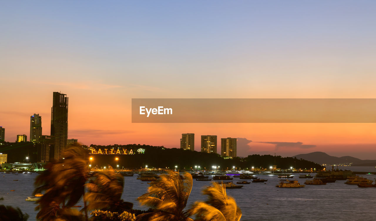 ILLUMINATED BUILDINGS BY SEA AGAINST SKY DURING SUNSET IN CITY