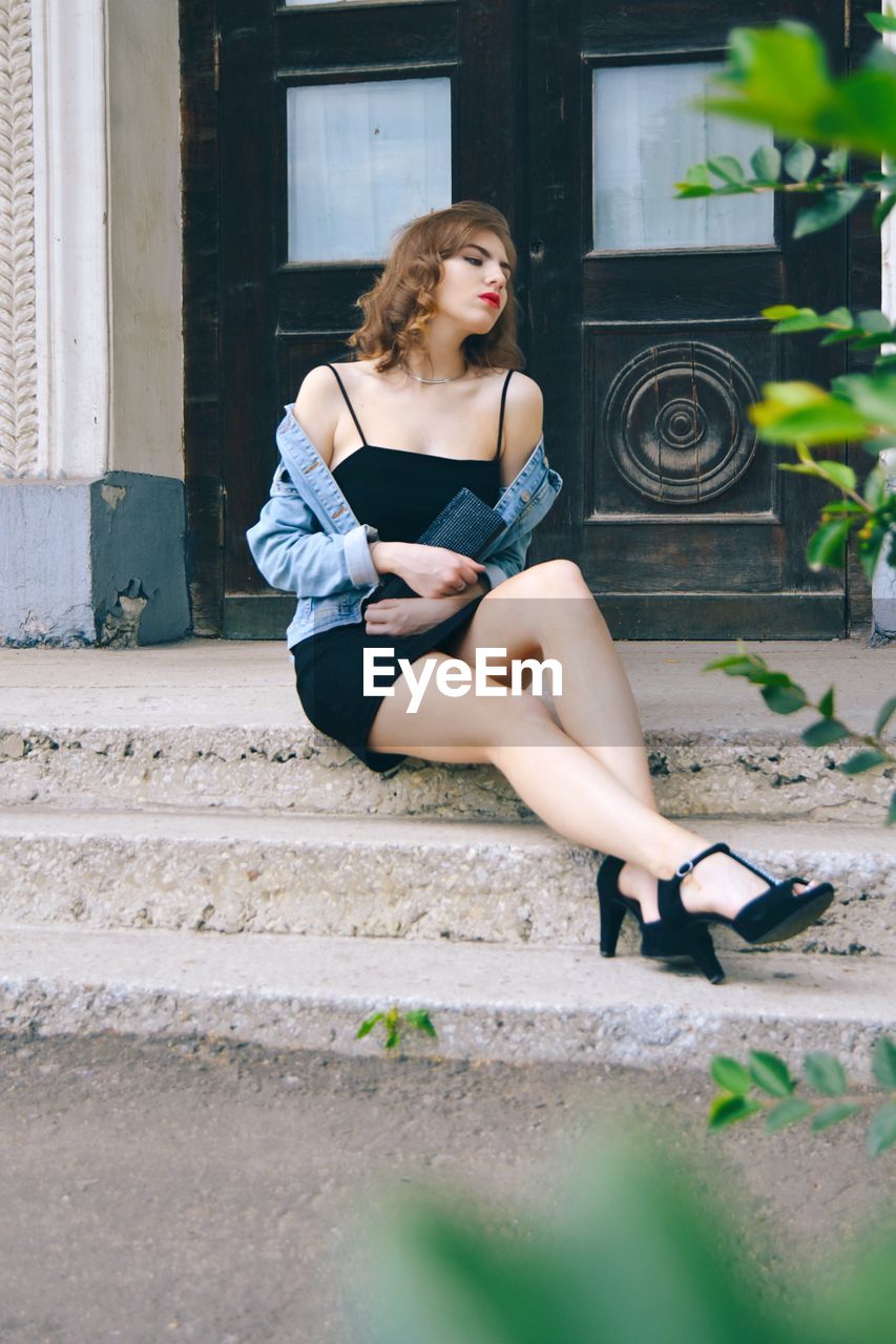 Full length of fashionable young woman wearing black dress sitting on steps