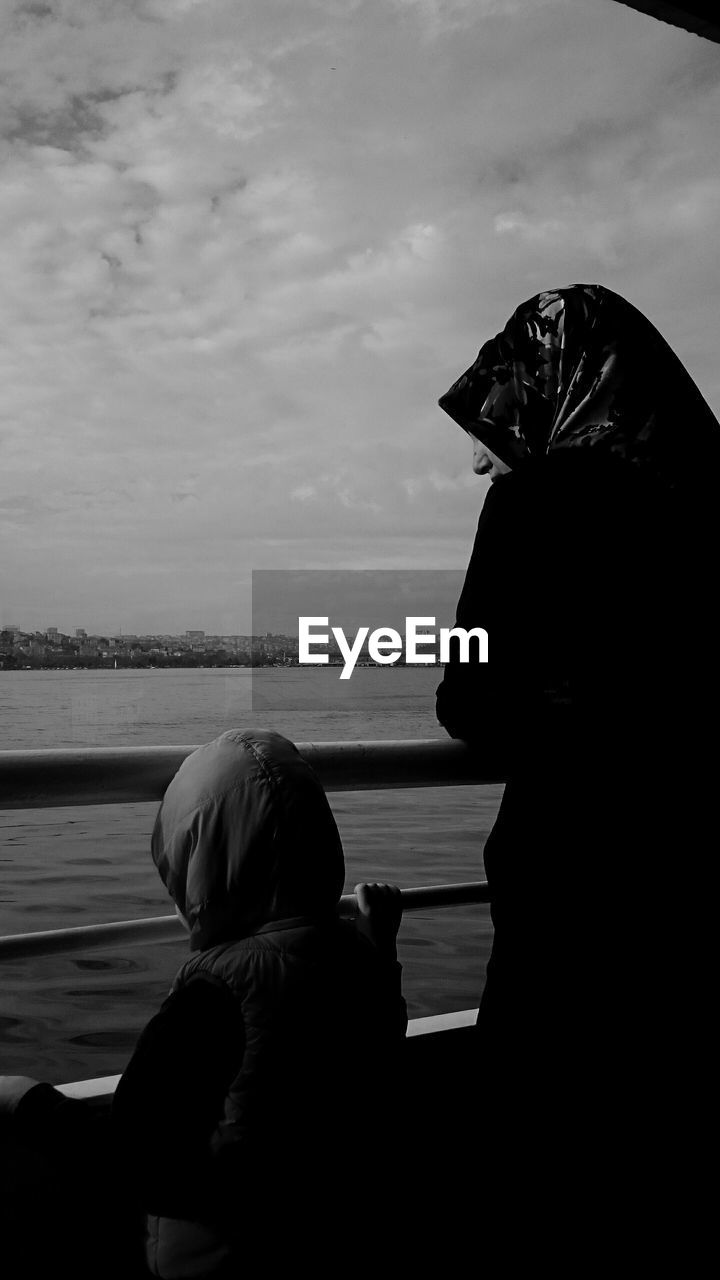 Rear view of mother and son standing by railing against cloudy sky