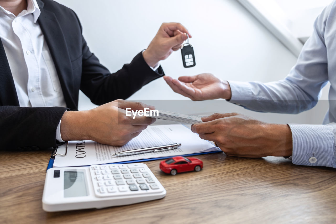 Agent giving car key to customer by papers and currency on table