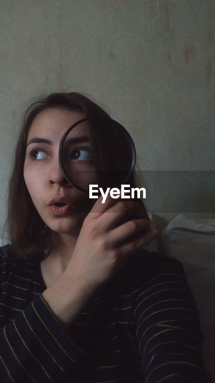 Close-up of young woman holding magnifying glass against wall