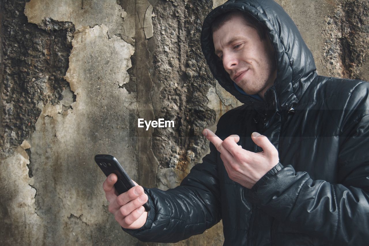 Man showing middle finger while holding mobile phone against broken wall