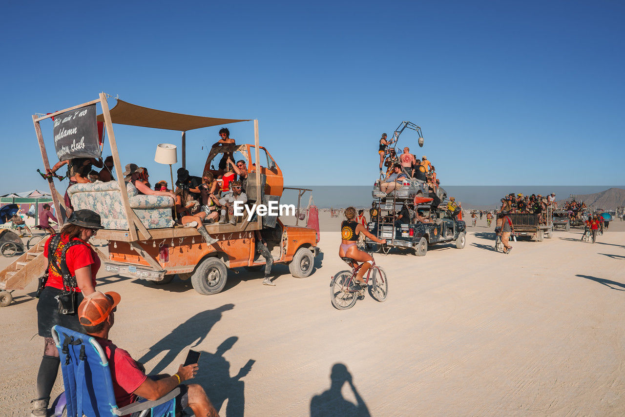 group of people, transportation, men, sky, adult, vehicle, nature, clear sky, beach, large group of people, women, bicycle, mode of transportation, crowd, sunny, land, sunlight, day, travel, sand, sports, leisure activity, outdoors, full length, blue, copy space, travel destinations, riding, sitting, motion, land vehicle, holiday