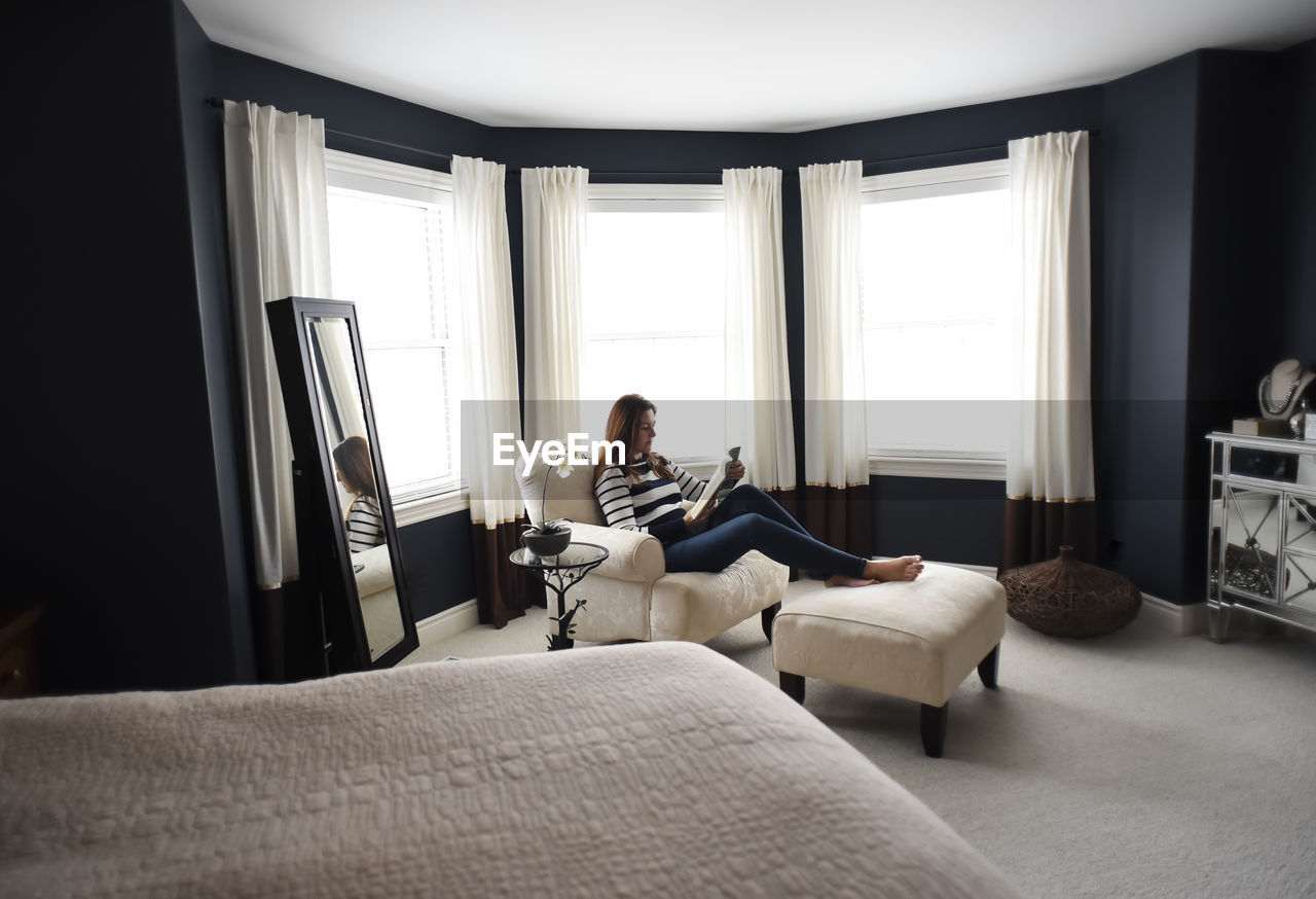 Woman sitting in chair on tablet in front of windows in a bedroom.