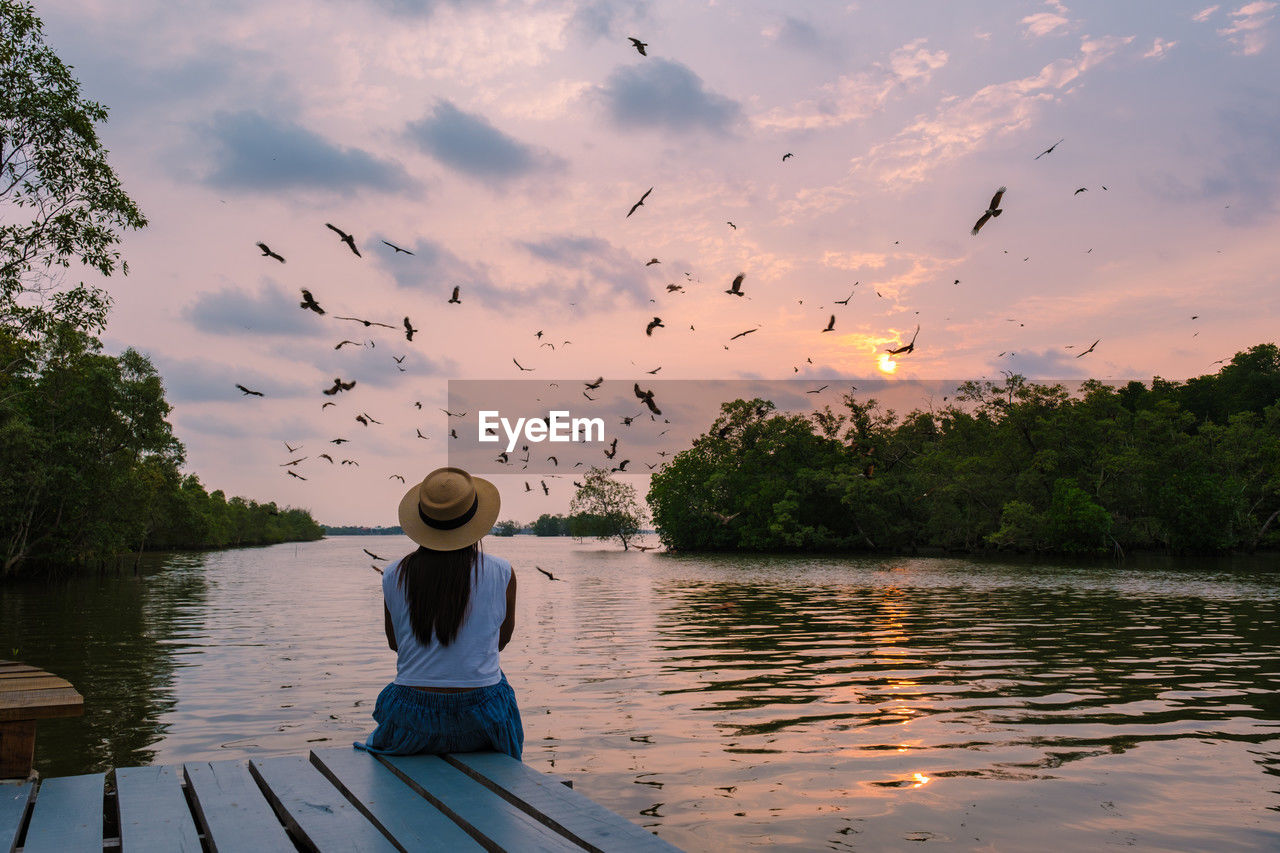 rear view of woman sitting on pier over lake against sky during sunset