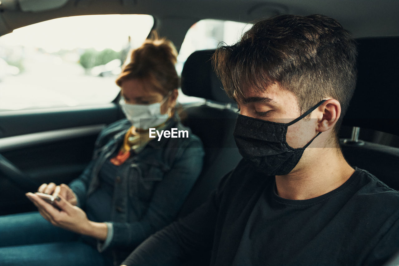 Young man and mature woman wearing mask sitting in car