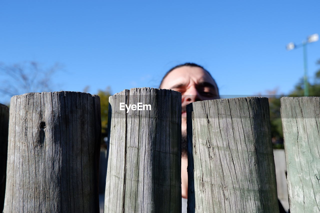 Portrait of man peeking over fence against sky