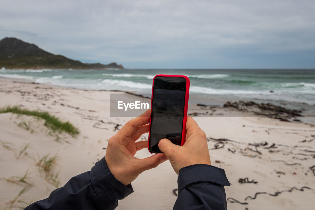 Hands of person holding mobile phone at beach against sky