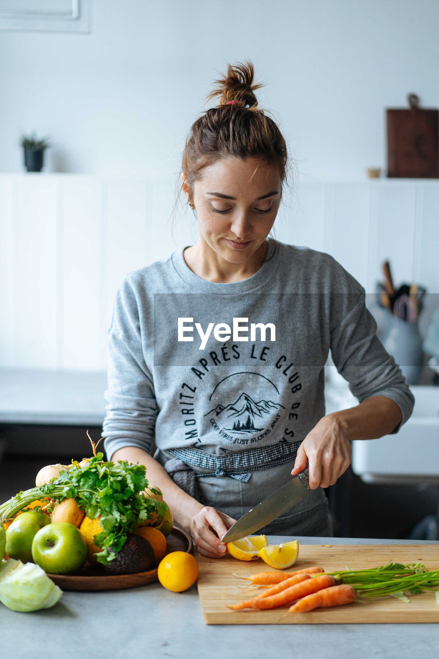 Adult lady slicing fresh lemon near ripe fruits and vegetables while standing in kitchen and preparing healthy dish for lunch