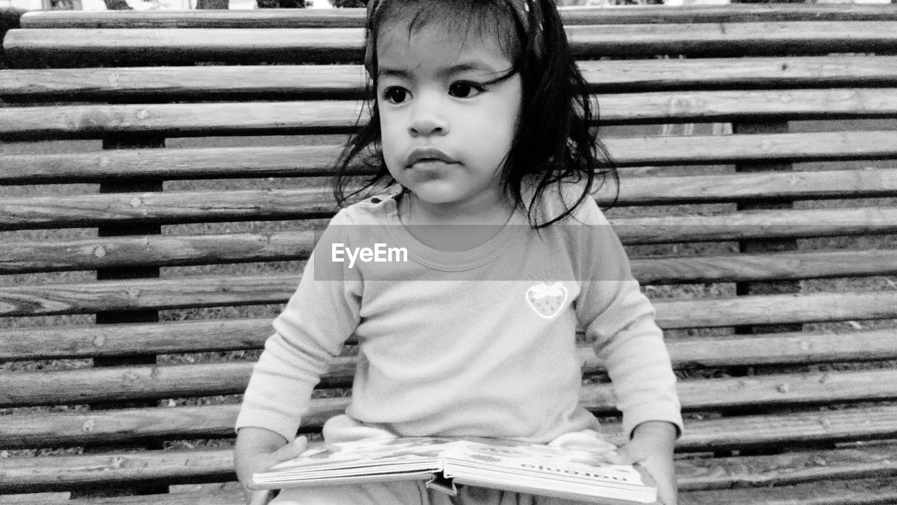 Cute baby girl holding book looking away while sitting on bench