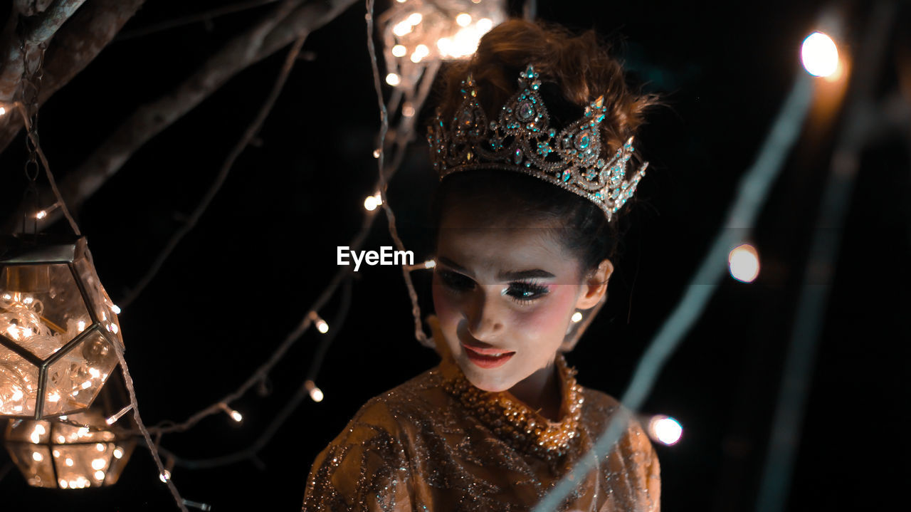 Close-up of beautiful model wearing crown amidst illuminated string lights at night