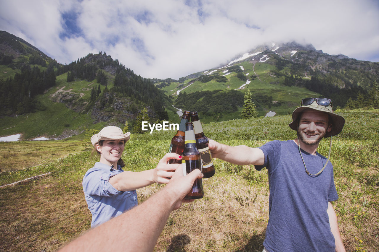 Happy friends toasting beer bottles on field against mountains and cloudy sky