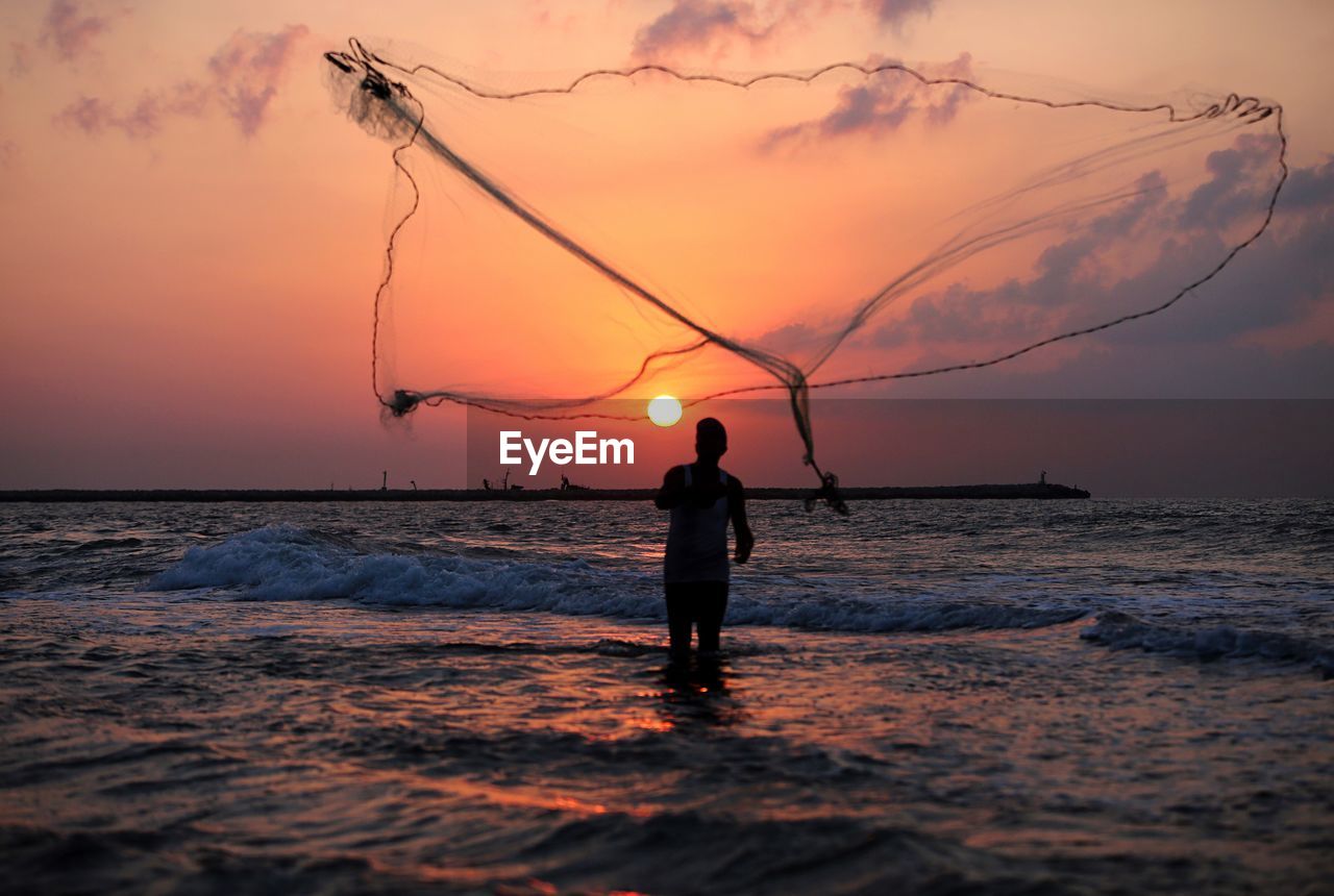 Silhouette man throwing fishing net in sea against sky during sunset