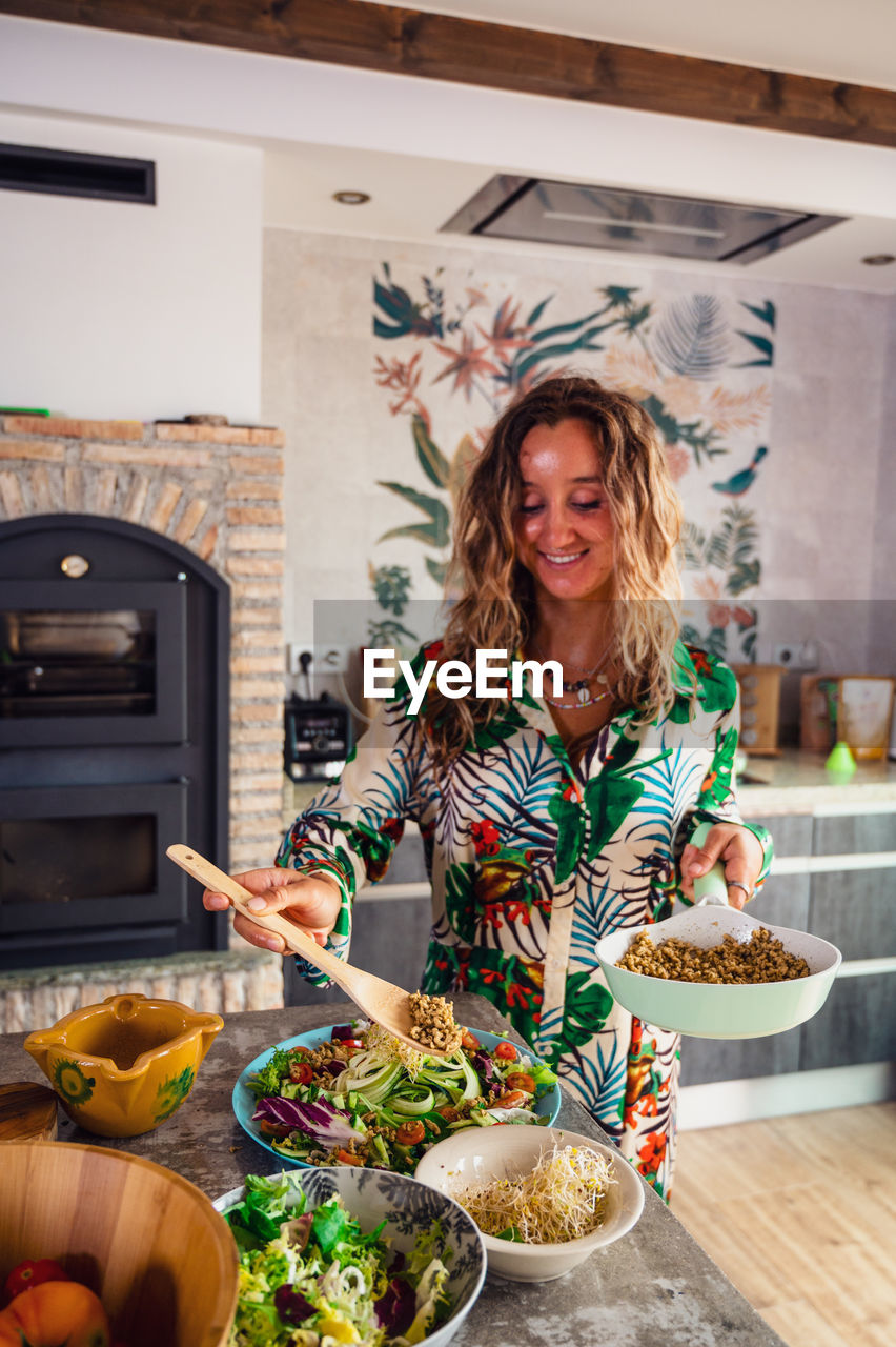 Female cook adding quinoa from frying pan in healthy salad with vegetables in light kitchen