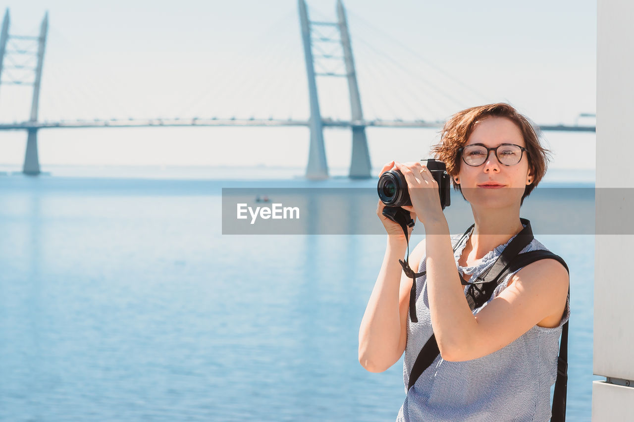 A tourist girl takes photos and smiles against the background of the bridge and the sea. 