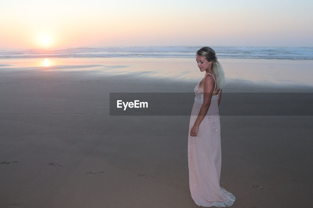 WOMAN STANDING ON BEACH AGAINST SEA DURING SUNSET