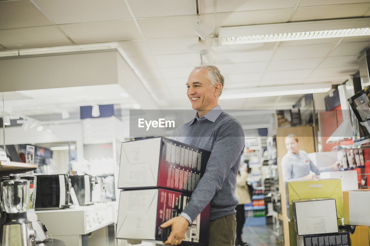 Smiling mature salesman with boxes walking in electronics store