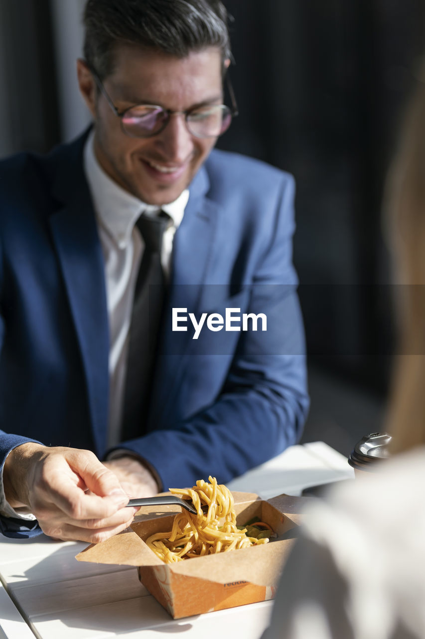 Businessman eating food while sitting with colleague in cafeteria at office