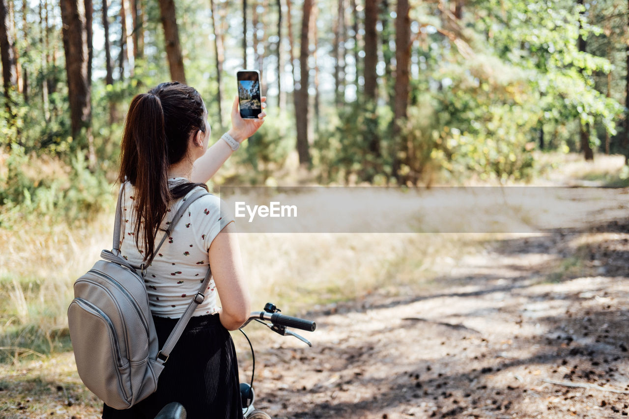 Young woman with backpack riding bike and taking photo on cell phone on pine forest background