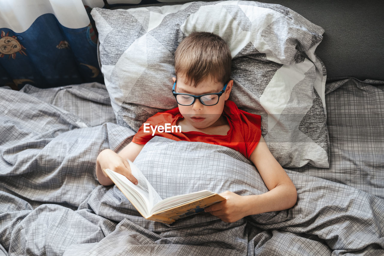 Boy with glasses lies on a bed under a blanket and reads a book, top view. 