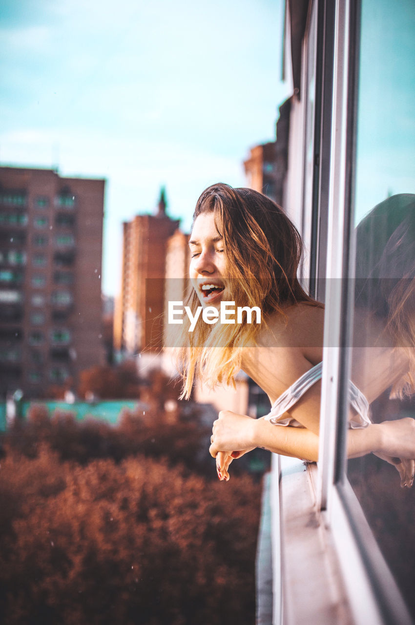 Side view of young woman leaning on window against buildings in city