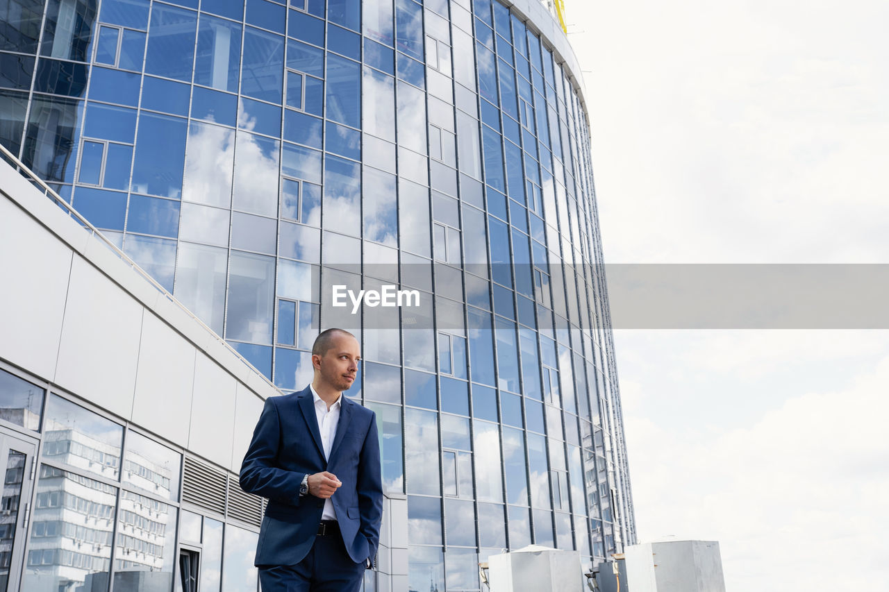 LOW ANGLE VIEW OF MAN STANDING BY MODERN BUILDING