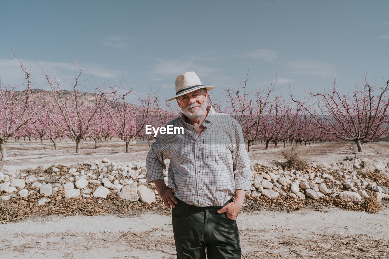 Content mature male farmer in hat looking at camera while standing near blossoming apricot trees in orchard