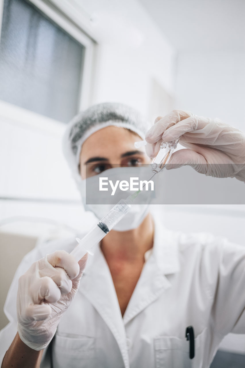 Vertical photo of a doctor with mask, cap and gloves filling a needle with anesthesia in an hospital room