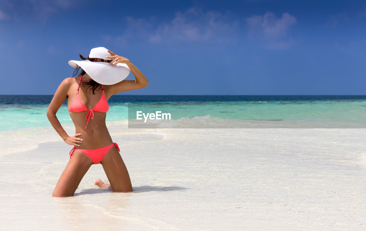 Young woman holding hat while kneeling on shore at beach against sky