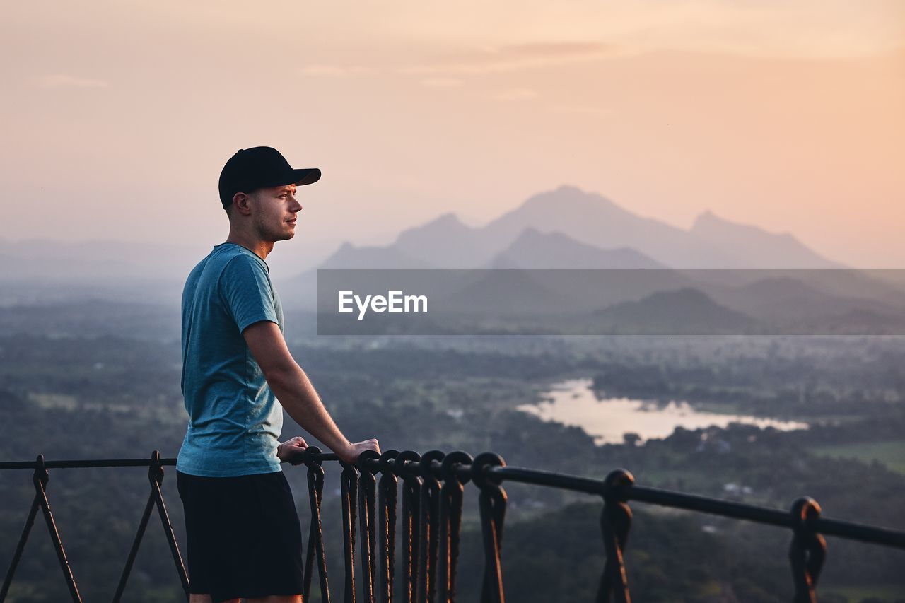 Side view of mid adult man looking at landscape while standing on mountain against sky during sunset