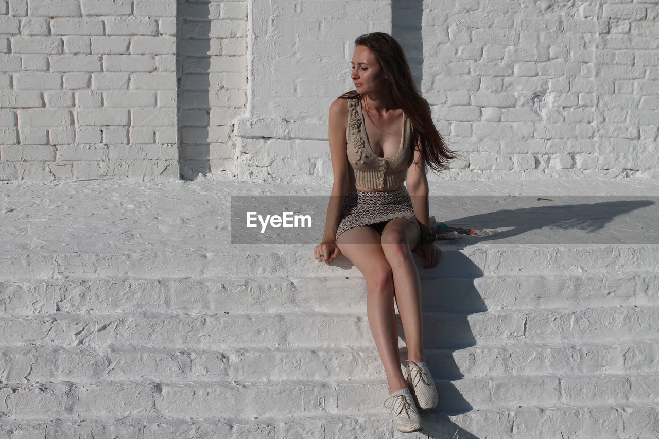 Full length of woman sitting against wall during sunny day