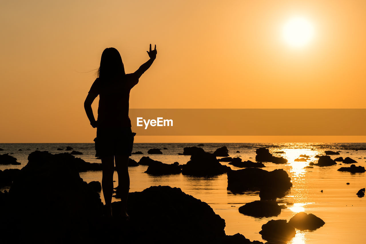 Silhouette woman gesturing while standing at beach against sky during sunset