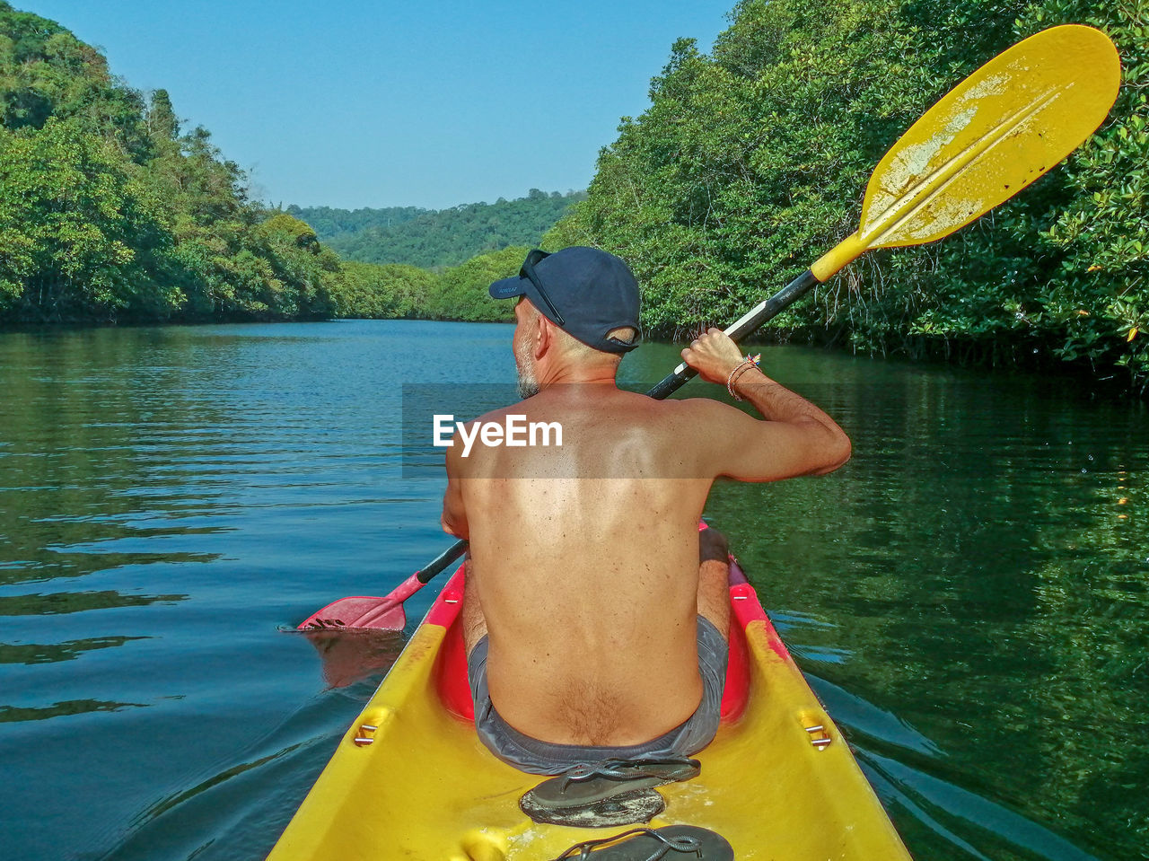 REAR VIEW OF SHIRTLESS MAN ON LAKE AGAINST PLANTS