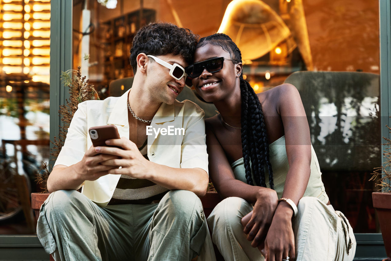 Happy black woman in stylish clothes and sunglasses smiling and leaning on boyfriend using smartphone while sitting against glass wall on city street