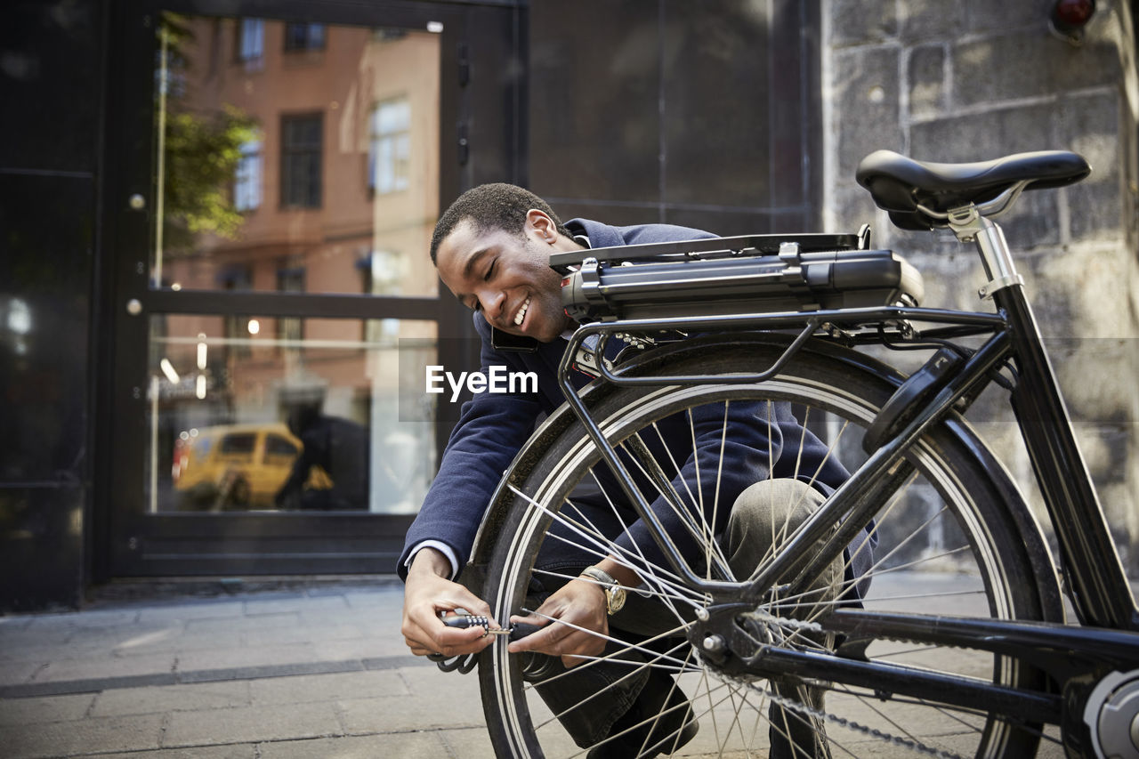 Smiling young male commuter locking electric bicycle while crouching against building in city