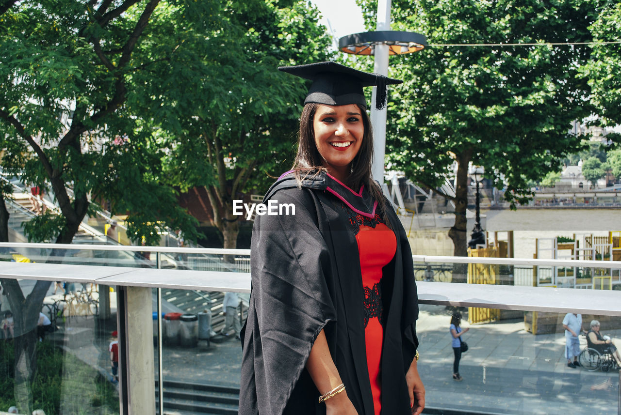 Portrait of smiling young woman wearing mortarboard against trees